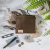 Image shows a Fernweh UK waxed cotton pouch, with the Fernweh logo etched in leather in the bottom left corner. The pouch sits on light coloured rocks on a light coloured background and is surrounded by a number of items that would fit inside the pouch such as snips, pens, keys, coins and rocks. .  