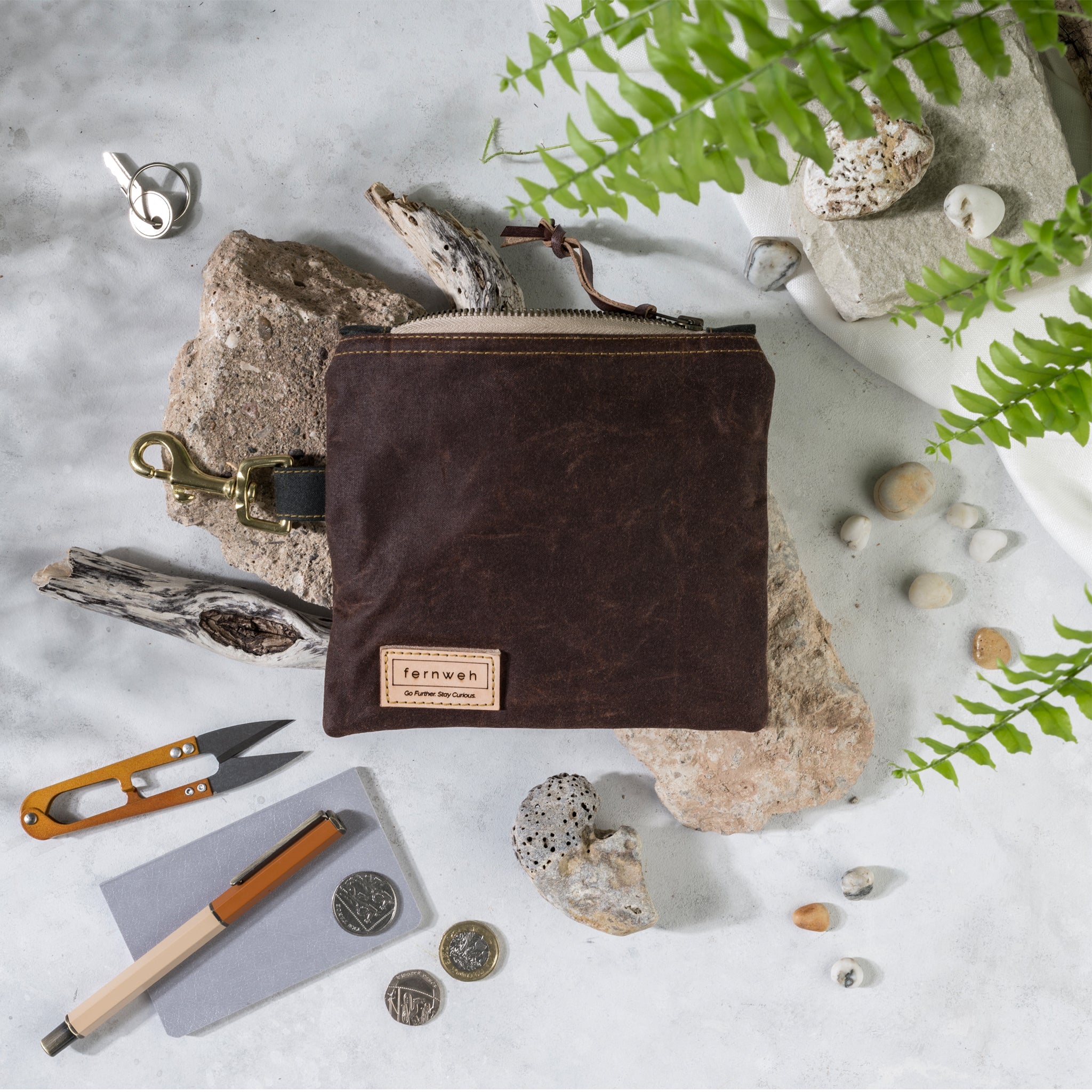 Image shows a waxed cotton canvas pouch in a rust colour, with a beige zip. The pouch sits on light coloured rocks on a white background. The zip is open. around the pouch are an array of items that would fit in the pouch, mustard snips, a mustard pen, coins, keys and small rocks