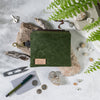 Image shows a moss green waxed cotton canvas pouch, sitting atop light coloured rocks. Surrounding the pouch are small items that can be stored in the pouch, such as keys, snips, pens and coins.