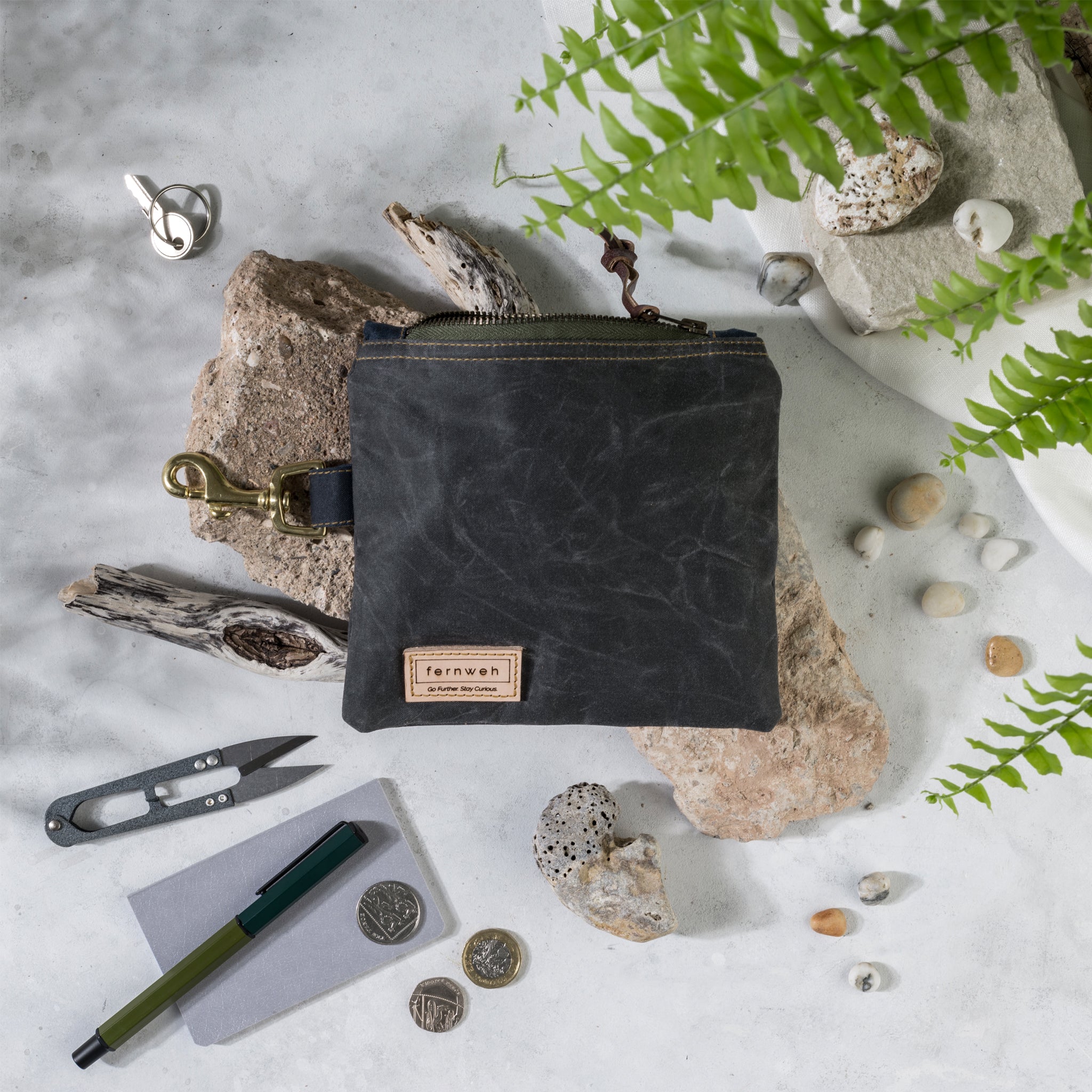 Image shows a waxed cotton pouch with a green zip, sitting on light coloured rocks. Around the pouch is an assortment of items that will fit inside the pouch, such as snips, pens, coins and small rocks