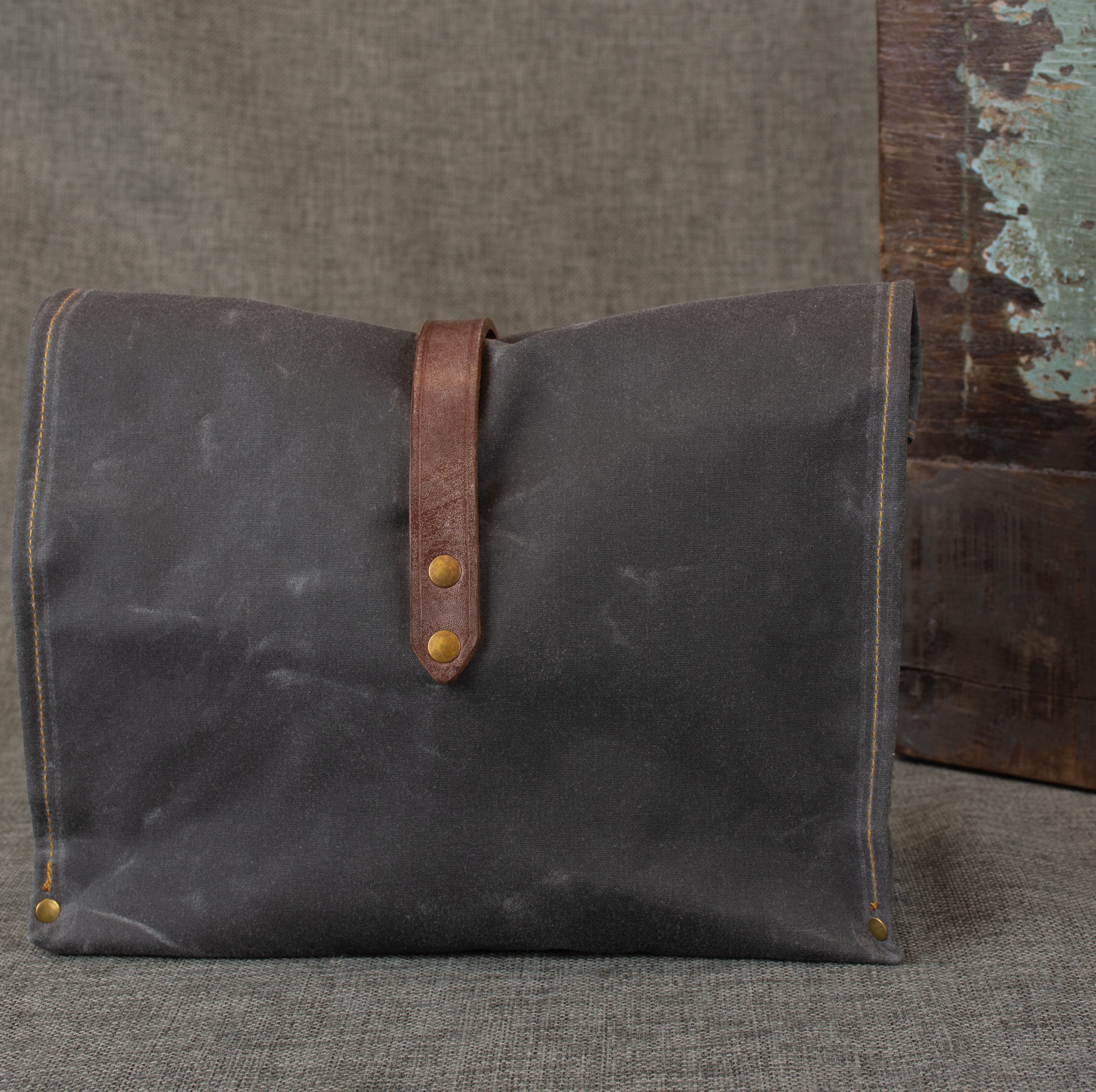 Image shows back view of a grey waxed cotton roll top bag with a brown oak bark tanned leather strap on a grey linen background.