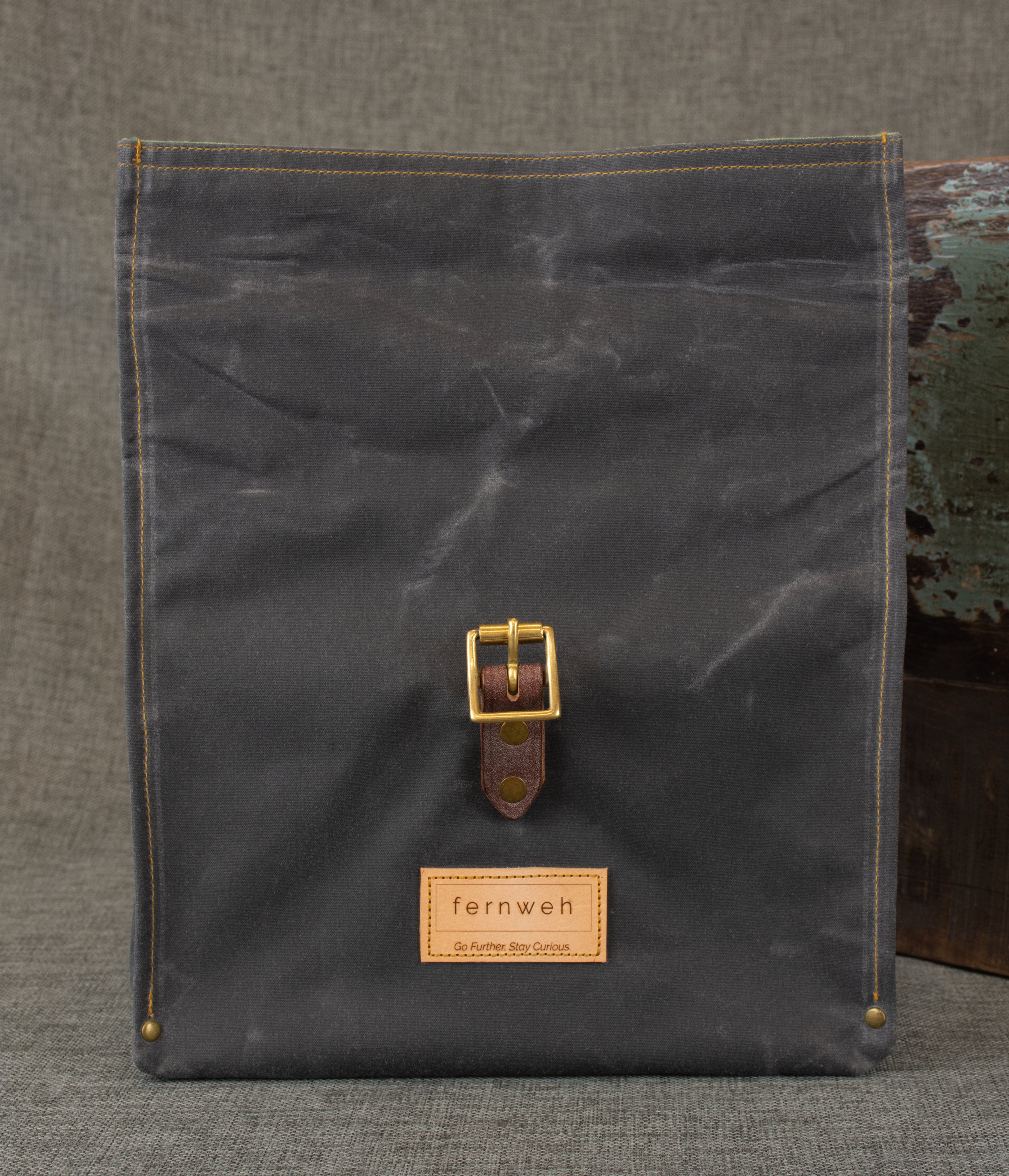 Image shows front, unrolled view of a grey waxed cotton roll top bag with a brown oak bark tanned leather strap on a grey linen background.